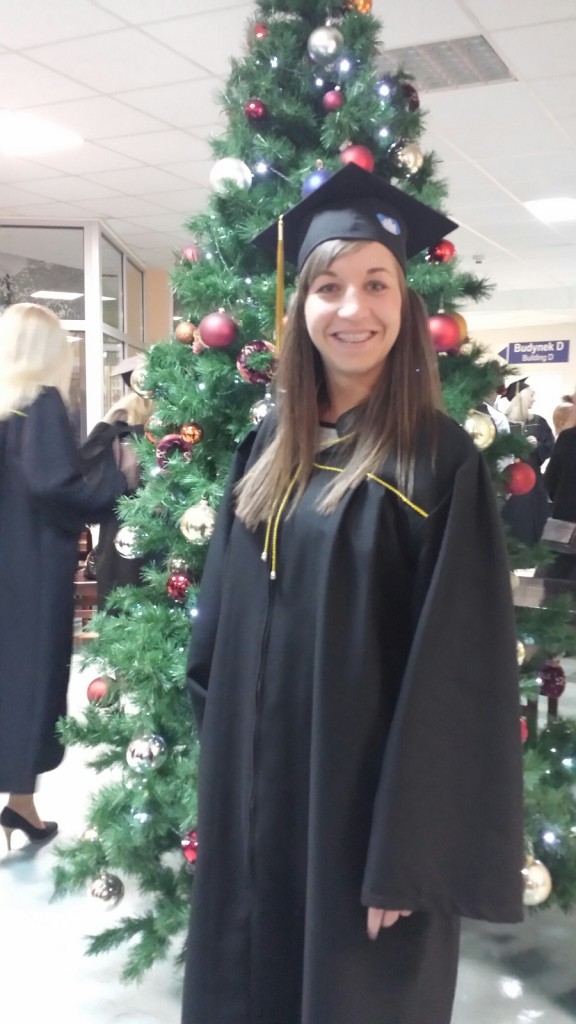 Young woman standing in graduation cap and gown before a Christmas tree.
